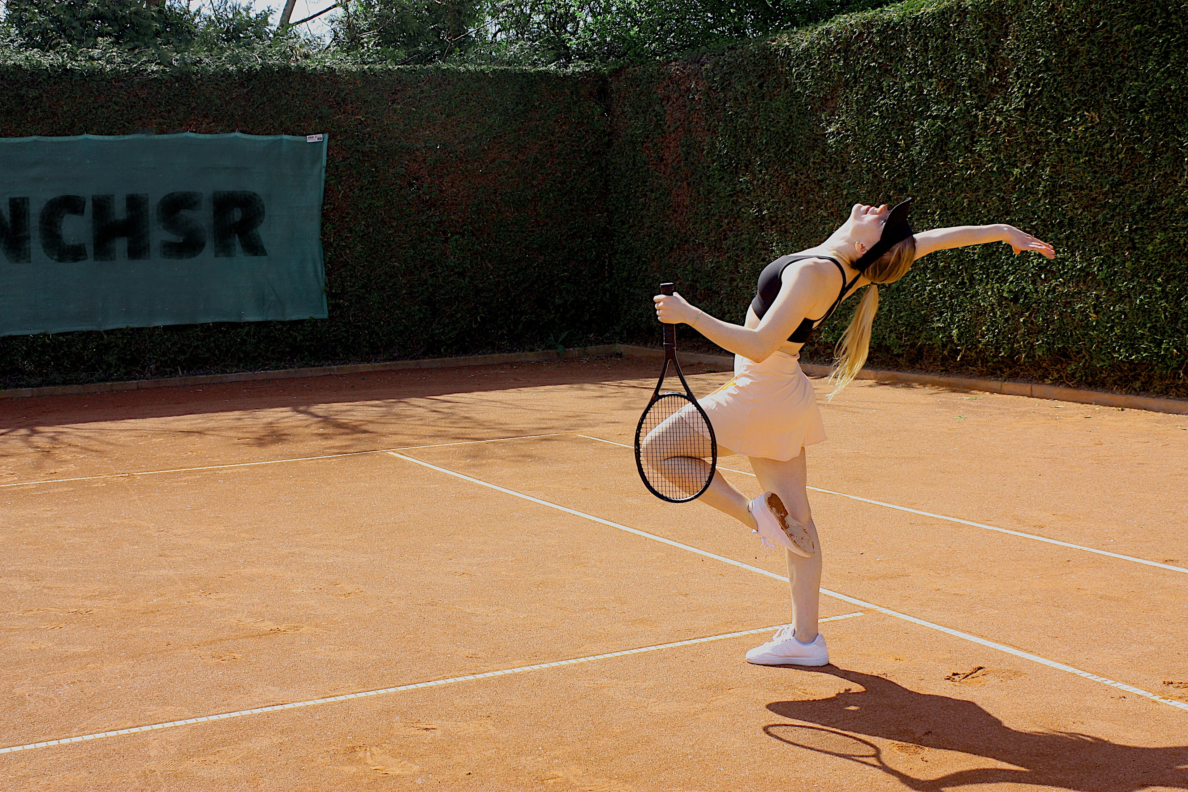 Das Bild zeigt eine Frau in einer coolen Tanzpose auf einem Tennisplatz mit einem Tennisschläger und sie trägt Tennischmuck.
